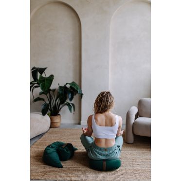 Kobieta siedzi tyłem w pozycji medytacyjnej na zielonej poduszce do relaksacji w jasnym wnętrzu.